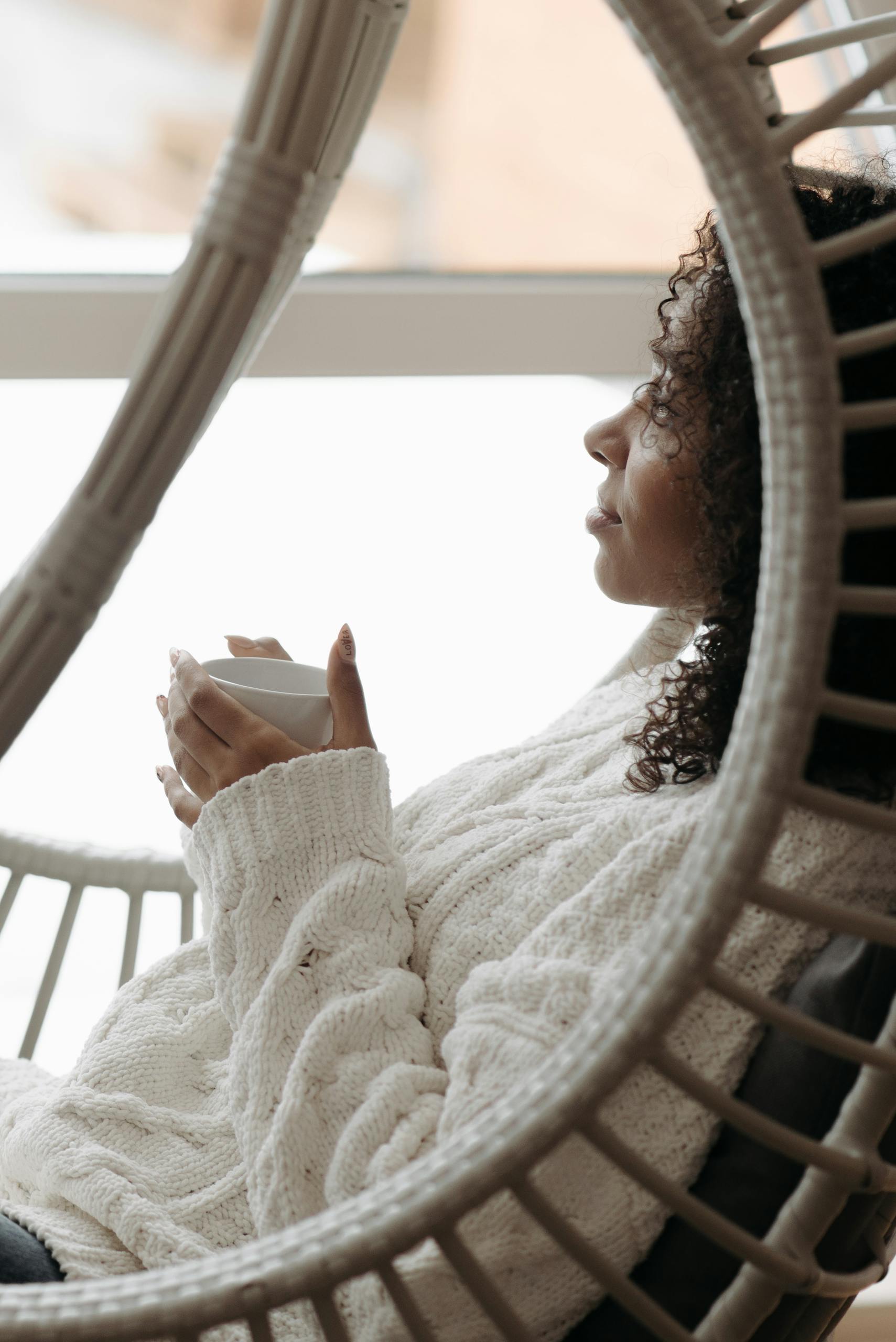 Peaceful woman with curly hair enjoys coffee in a stylish indoor setting, captured in profile view.