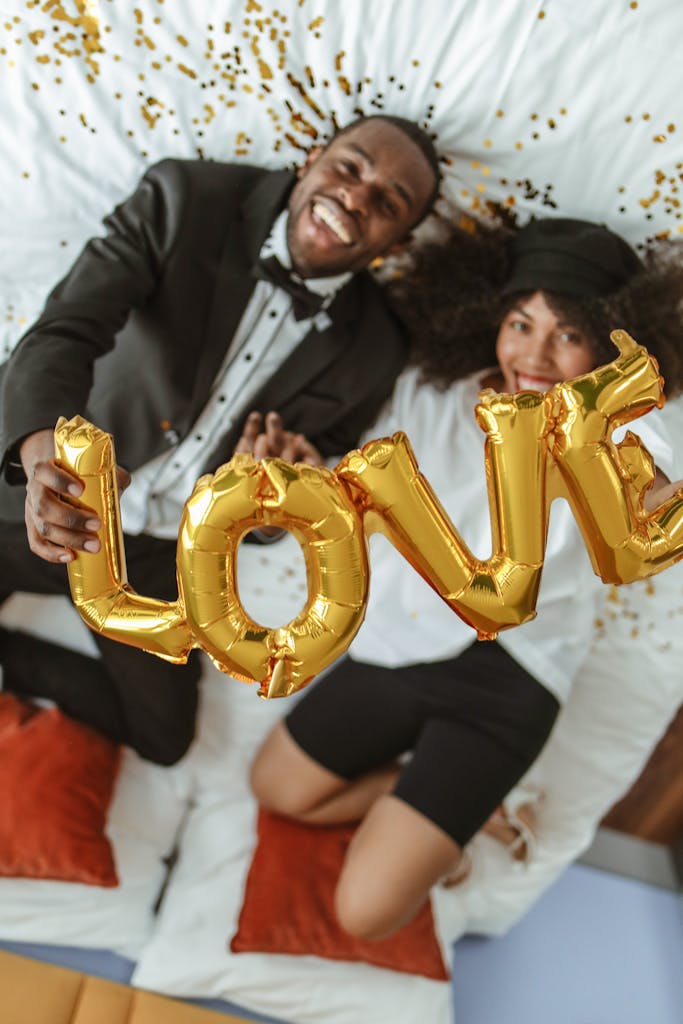 Smiling couple in formal attire holding love balloons on a bed with confetti, capturing a joyful and romantic moment.